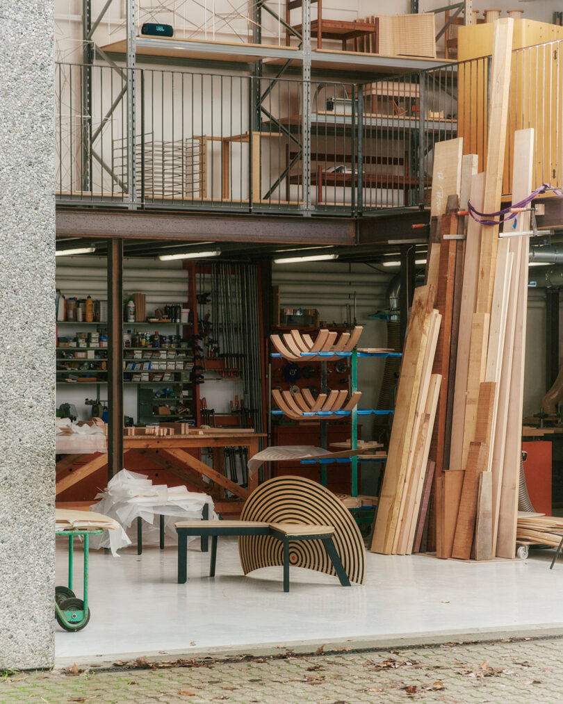 A woodworking studio with stacked lumber, shelves of tools and supplies, and finished wooden furniture pieces visible in the foreground.