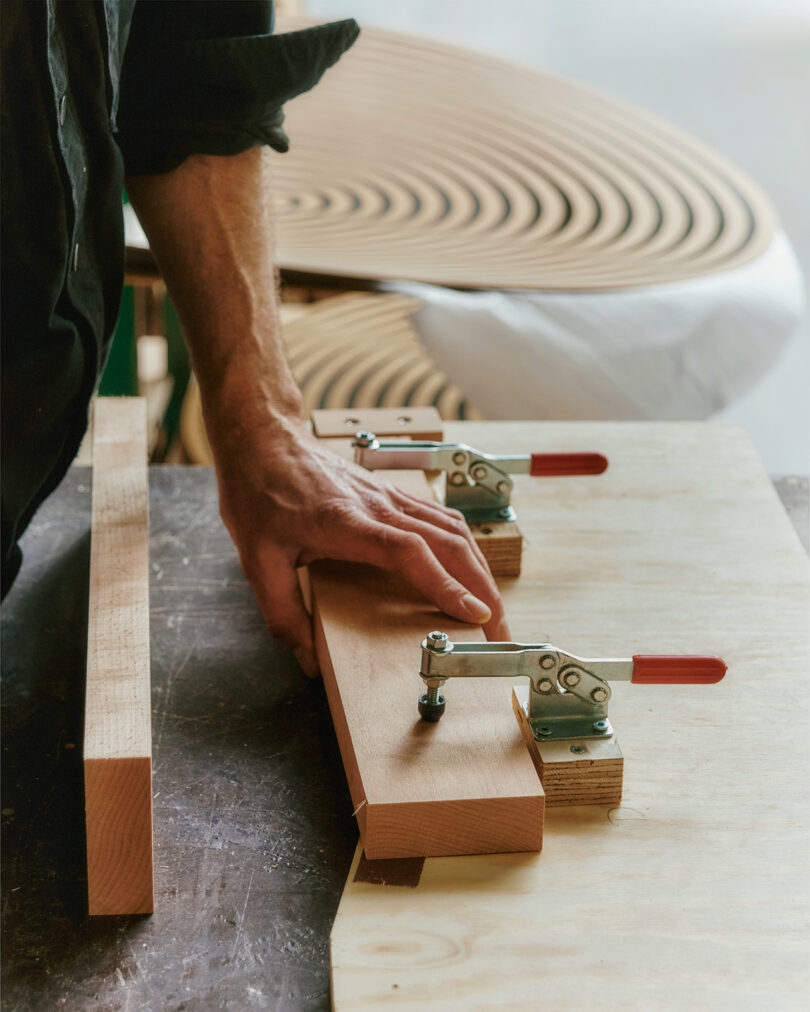 A person’s hand adjusts a piece of curved wood secured by two toggle clamps on a workbench, with additional curved wood sections in the background.