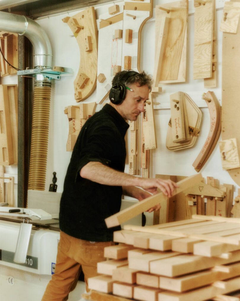 A man wearing headphones works with wooden boards in a workshop, surrounded by various woodworking tools and templates on the wall.