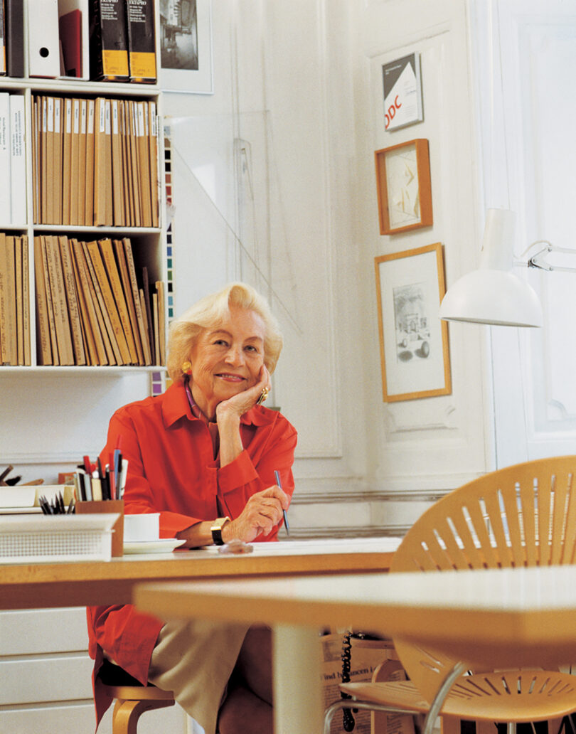 Nanna Ditzel in a red shirt sits at a desk in a bright office, smiling, with shelves of files and framed artwork on the wall behind her.