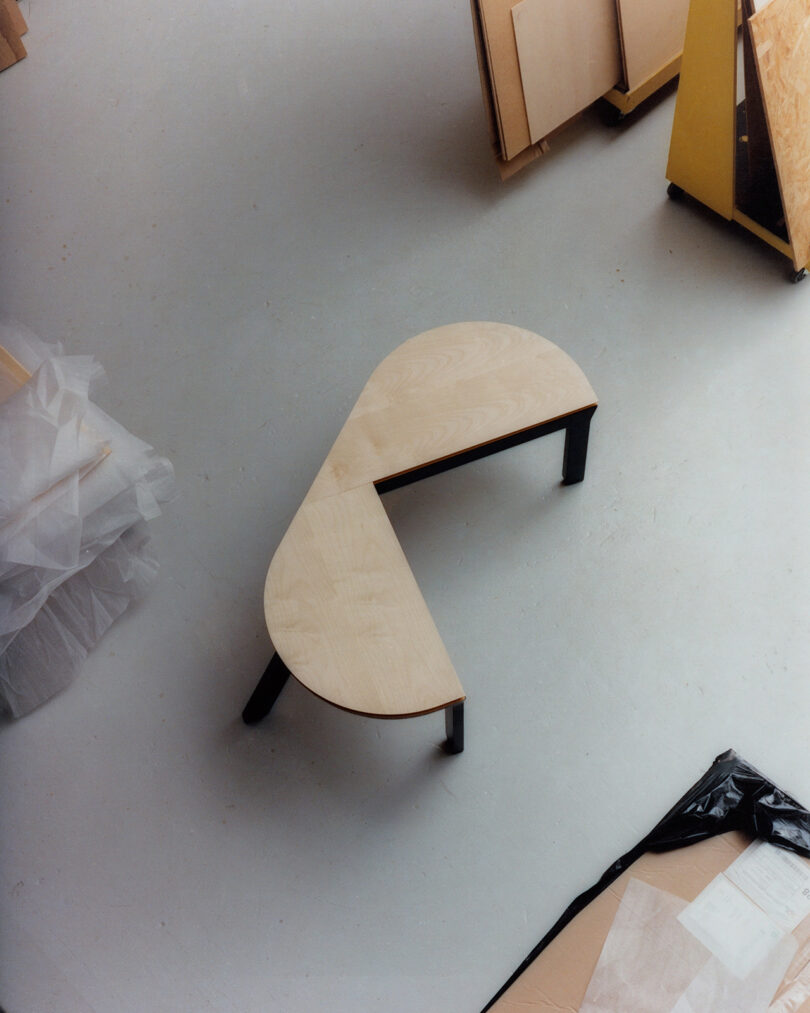 A wooden, semi-circular table with a triangular cutout stands on three black legs in a workshop with scattered boards and plastic sheets.