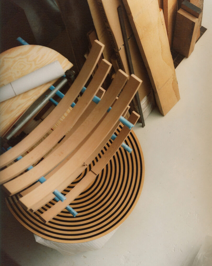 A stack of curved wooden slats and a round, layered wooden piece with concentric circles, surrounded by various wood planks on a light floor.