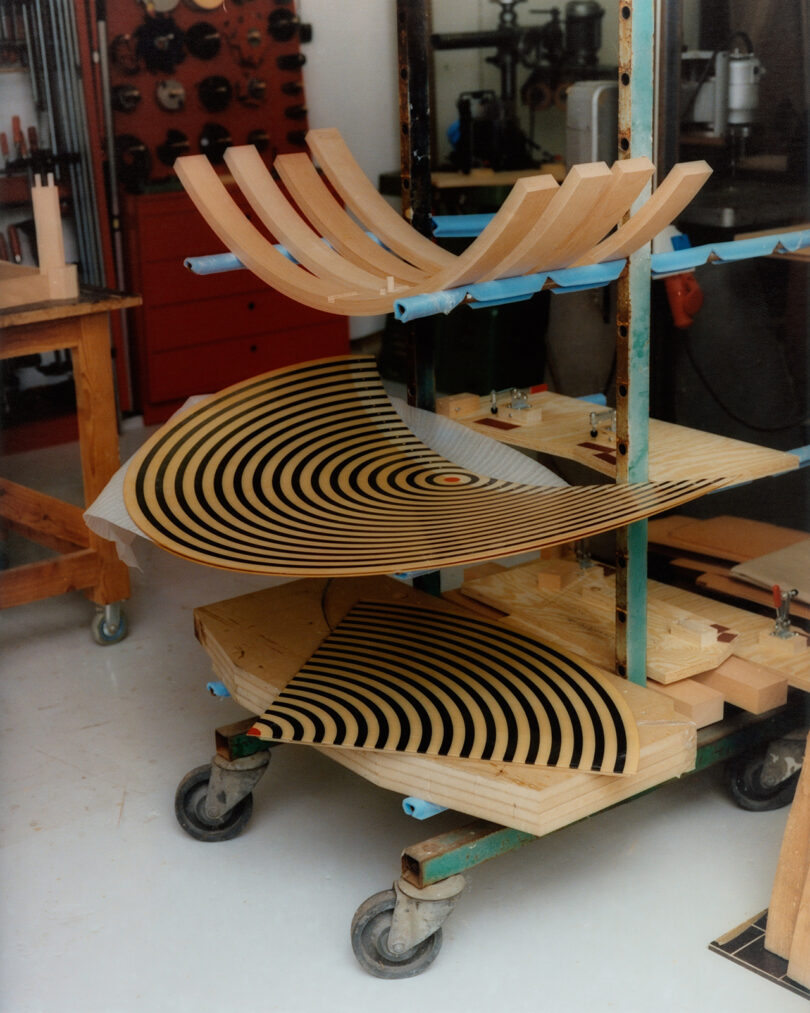 A mobile cart holds curved wooden pieces and two semi-circular panels with black concentric circles, set in a workshop with tools and machinery in the background.