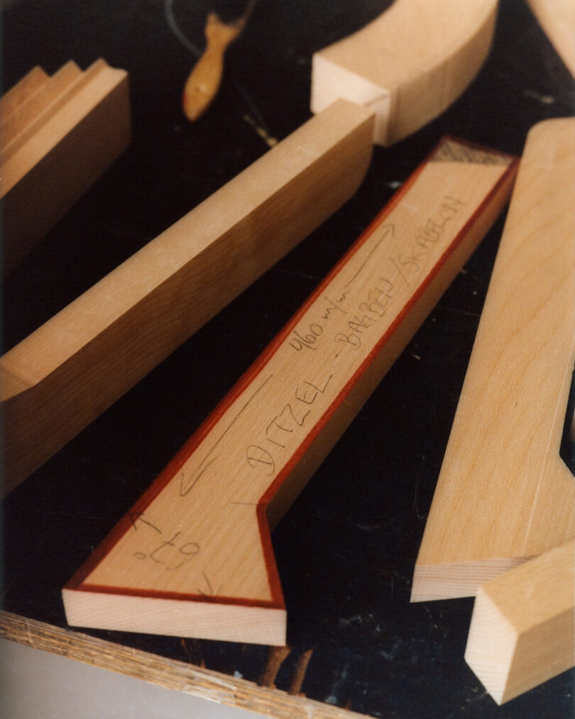 Several unfinished wooden pieces, including an arrow-shaped board with handwritten text and measurements, are arranged on a dark surface.