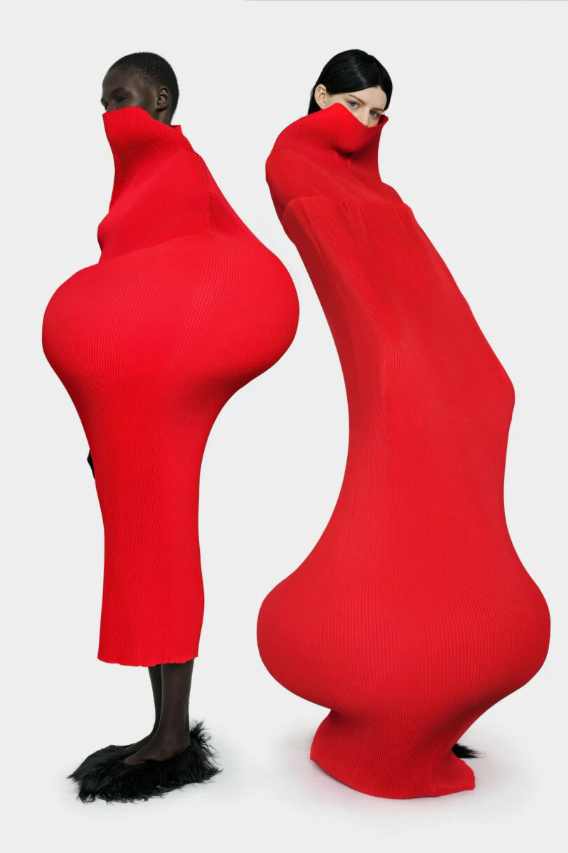 Two models wear exaggerated red dresses, fit in shape, with extreme hourglass shapes. A dress covers the lower face; Both outfits distort the silhouette of the natural body on a simple background.