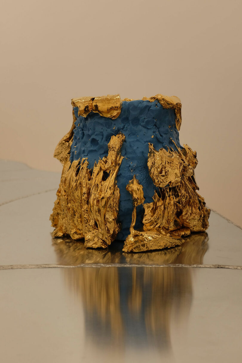 A blue ceramic vessel with textured, irregular gold accents on the top and base, displayed on a reflective metal surface on a neutral background.