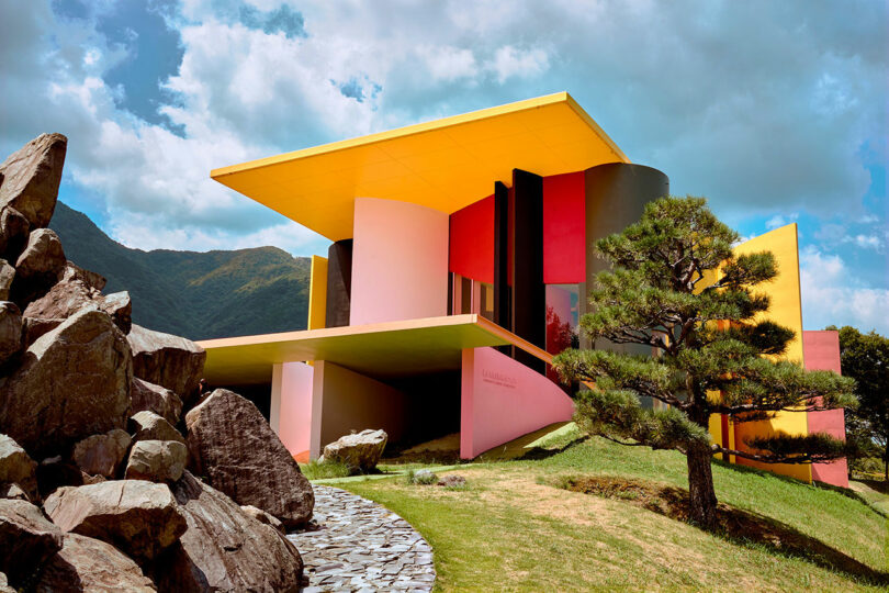 Modern house, with bold geometric shapes and bright colors, including yellow, pink, black and red, placed in a courtyard with rocks and a tree under a partially cloudy sky.