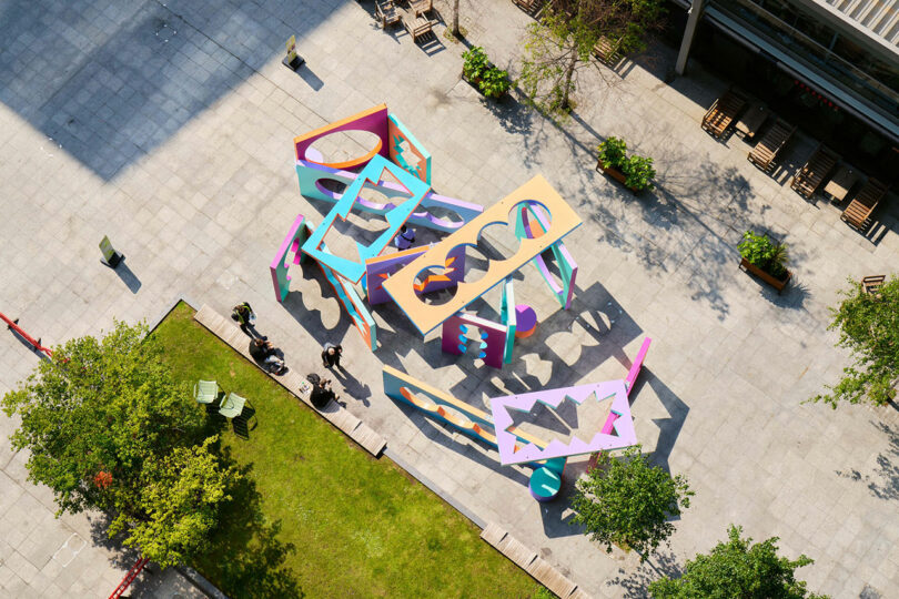 Aerial view of an open -air colored geometric art installation on a paved market, surrounded by trees, grass and people nearby.