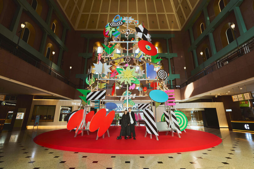 A large, colorful abstract sculpture with geometric shapes, sits on a red carpet in the center of a spacious atrium of the mall, with two people posing in front.