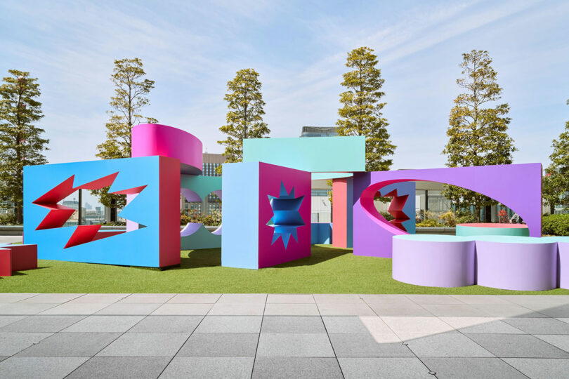 The colored geometric sculptures with star -shaped cuts are displayed on a grassy area, trees and a building in a clear sky.