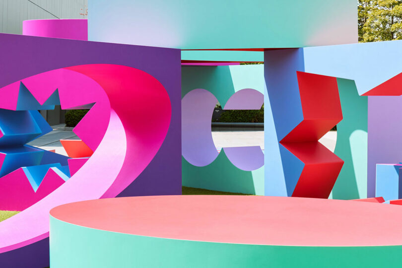 Colored geometric sculptures, with circular, rectangular and jammed cuts, arranged outdoors, with vibrant pink, purple, blue, green and red.