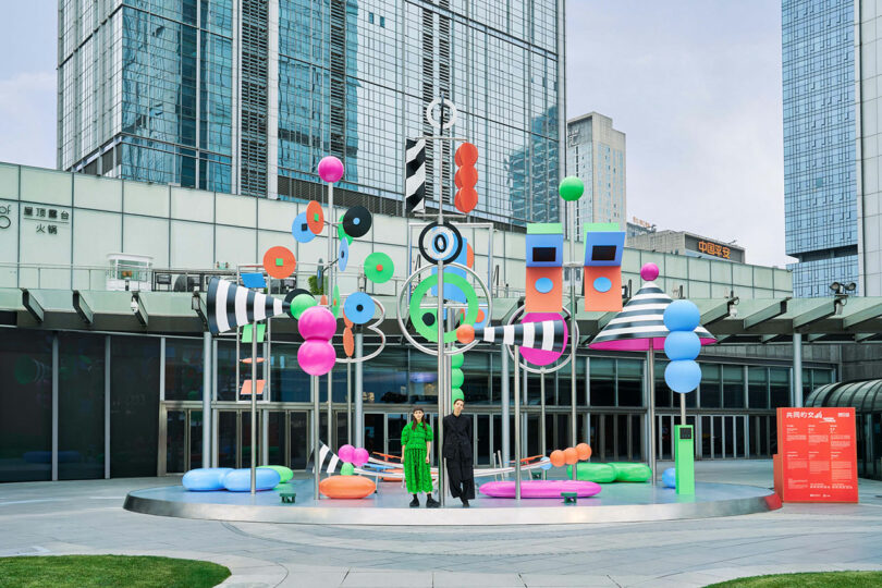 Two people stand in front of a colored art installation, abstract outdoors, with geometric shapes and large buildings in the background.