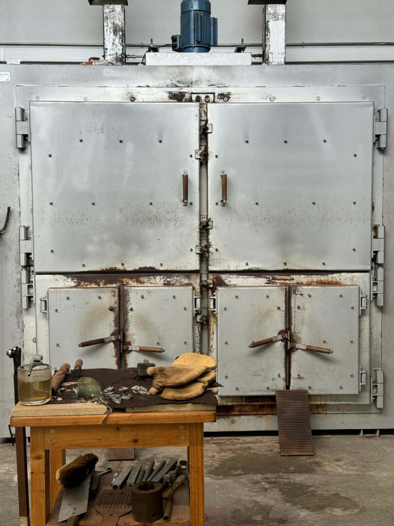 A large industrial metal furnace designed by Renaud Defrancesco stands behind a workbench holding tools, protective gloves, a glass jar, and metal scraps.