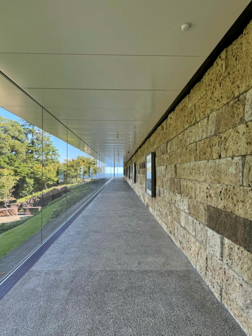 A modern corridor designed by Renaud Defrancesco features a glass wall on the left showcasing green trees, while a stone wall with framed displays lines the right. The corridor leads invitingly toward natural light.