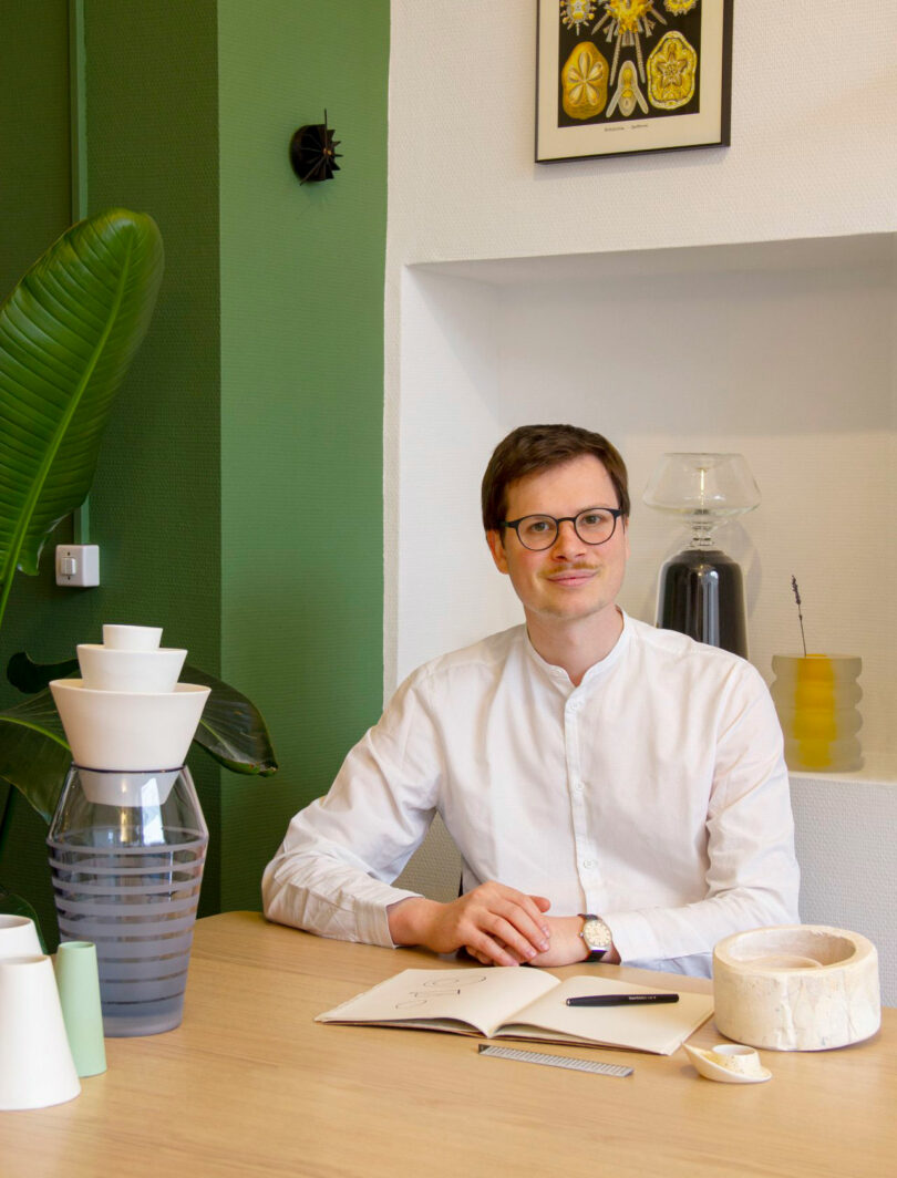Renaud Defrancesco, a man in glasses and a white shirt, sits at a desk with notebooks, ceramic items, and decorative objects in a modern, green-accented room.