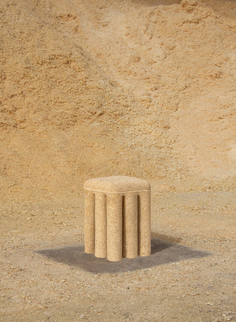 A small, cylindrical stool with four vertical legs, designed by Renaud Defrancesco and made of a light, sandy material, is placed on a ground covered with fine, beige sawdust.