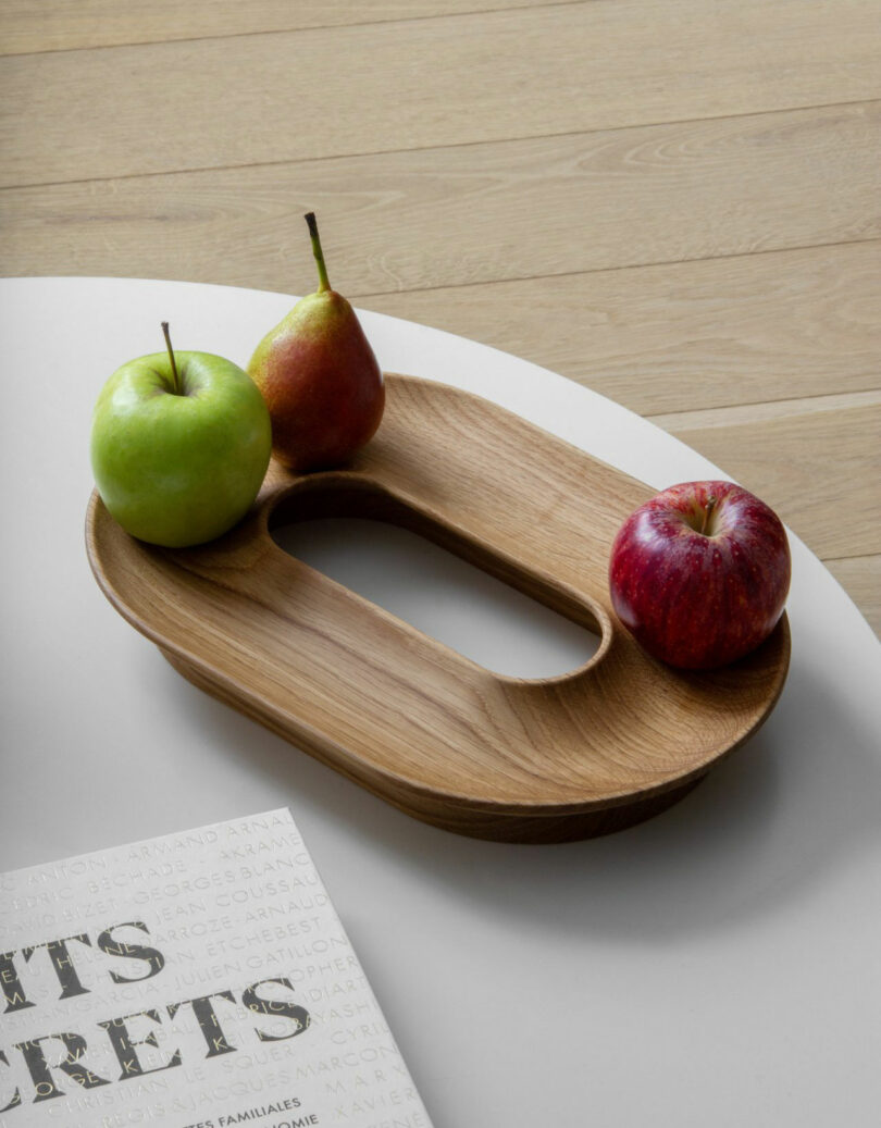 A wooden tray by Renaud Defrancesco with a cut-out center holds a green apple, a red apple, and a pear on a white table next to a closed book.