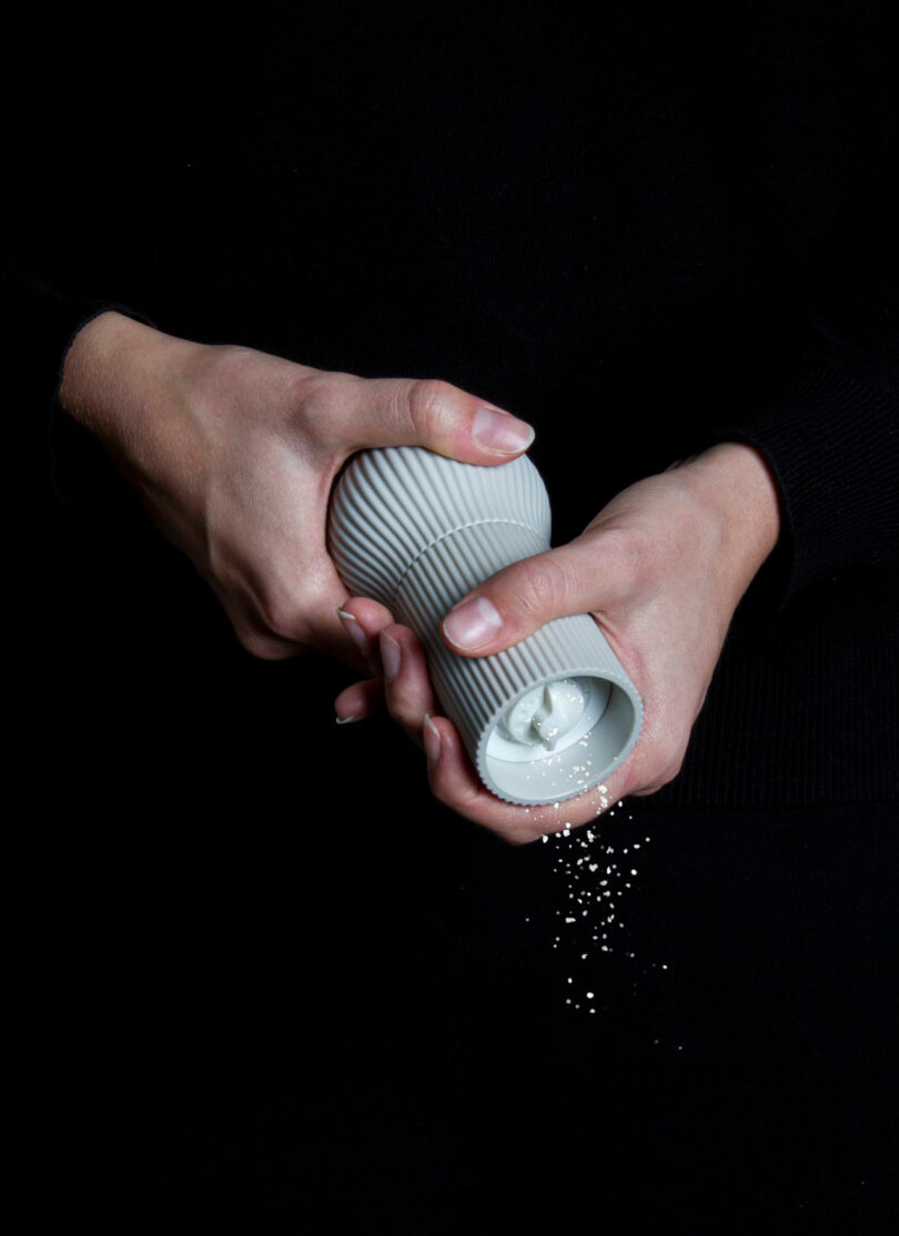 A person holding and twisting a white ribbed Renaud Defrancesco pepper grinder, dispensing ground pepper onto a black surface.