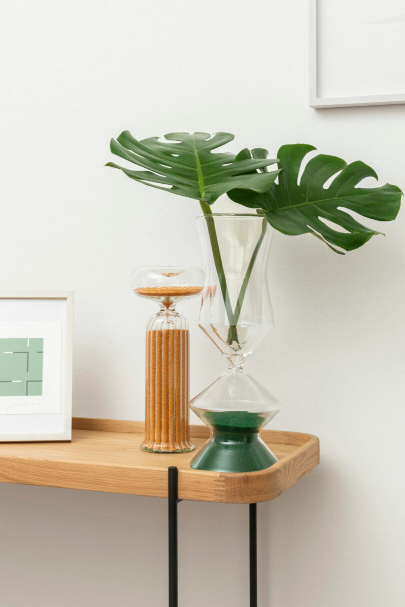 A wooden shelf displays a framed abstract print by Renaud Defrancesco, a clear vase with green monstera leaves, a glass vessel with sand, and a green sand hourglass.