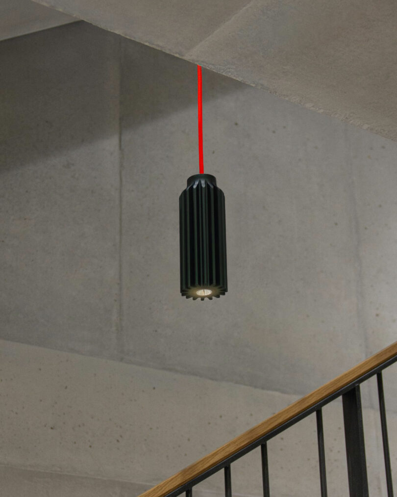 A black pendant light by Renaud Defrancesco with a red cord hangs from a concrete ceiling above a staircase featuring a wooden handrail.