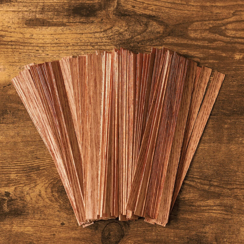 Several thin, reddish-brown wooden veneer strips are fanned out on a wooden surface, showcasing the craftsmanship reminiscent of Sam Sacks’ attention to detail.