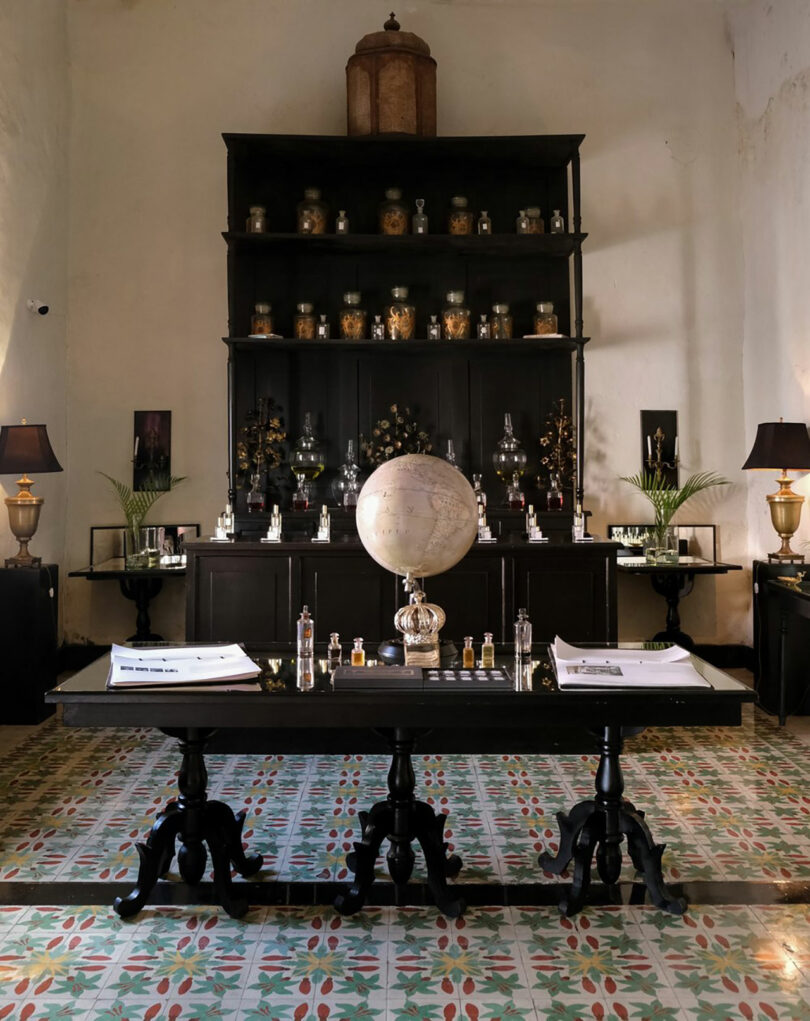 A vintage study room with a large globe on a wooden table, glass bottles, books, and a cabinet filled with jars and decorative items against the back wall—a space inspired by the eclectic charm of Sam Sacks.