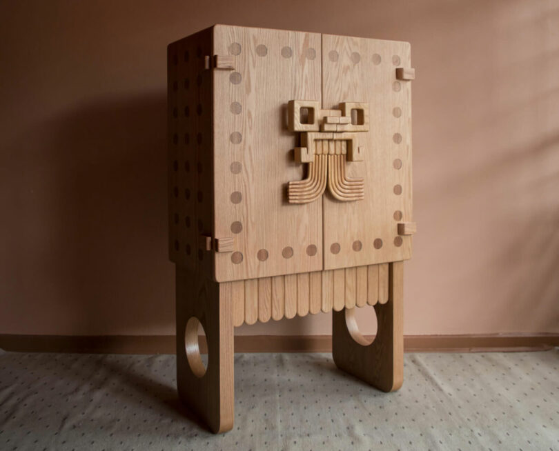 A light wood cabinet inspired by Sam Sacks, featuring dotted patterns, decorative carvings on the doors, and two rectangular legs with circular cutouts, positioned on a carpeted floor against a plain wall.