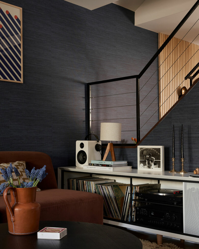 Modern living room styled by Sam Sacks, featuring a brown chair, record player, speakers, vinyl records, a table lamp, decorative accents, and stairs with a sleek metal railing in the background.
