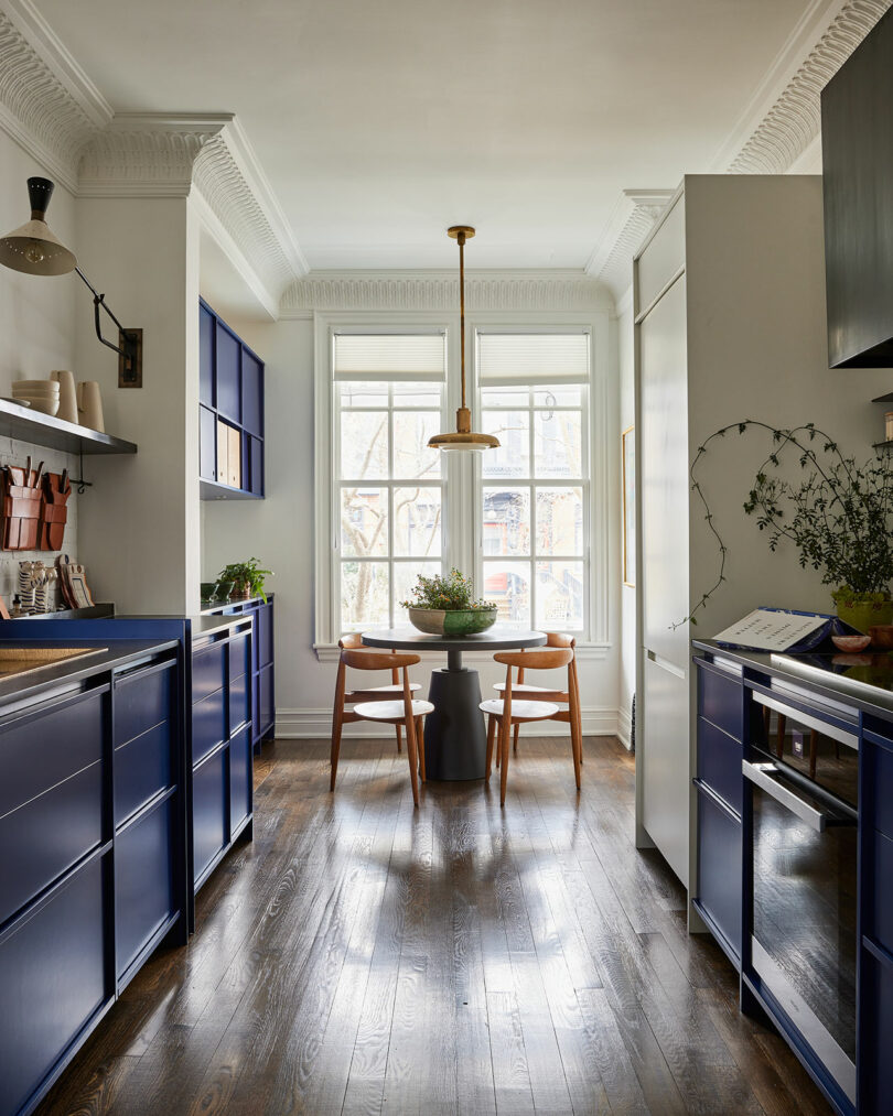 A modern kitchen by Sam Sacks with dark blue cabinets, wooden floor, a round dining table with four chairs, large window, and brass light fixture.