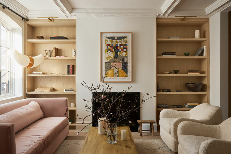 A modern living room inspired by Sam Sacks features a pink sofa, cream armchair, wooden coffee table, wall art above a fireplace, and built-in shelves holding books and decor items.