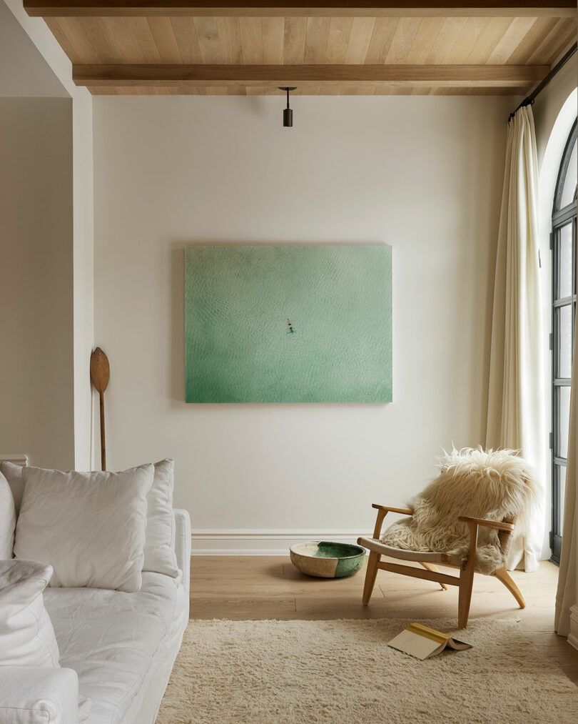 A minimalist living room designed by Sam Sacks features a white sofa, a wooden chair with a sheepskin throw, a large green abstract painting, and a bowl on a wood floor near an arched window.