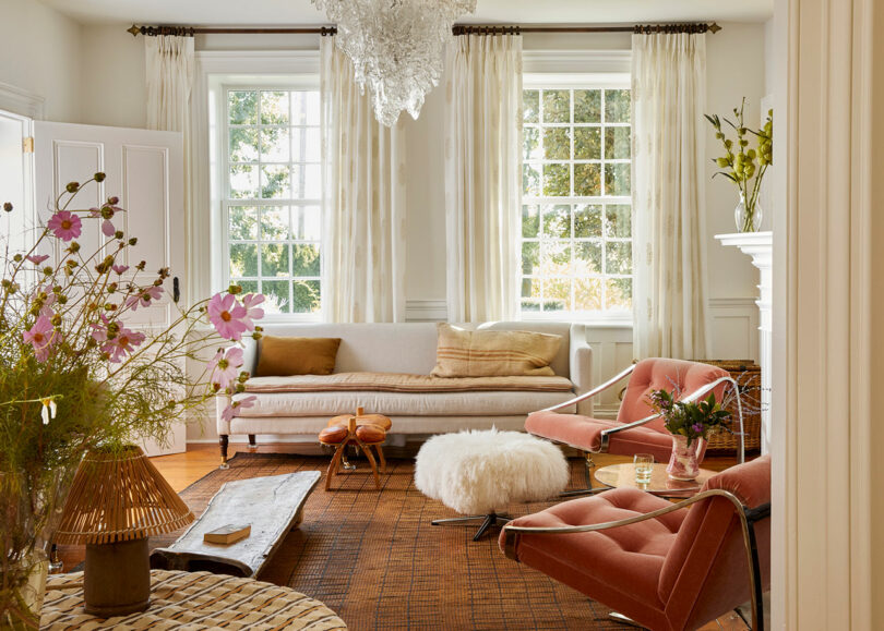 A bright living room by Sam Sacks with large windows, a white sofa, pink chairs, a fluffy round ottoman, and modern decor—including a chandelier and vases with flowers.