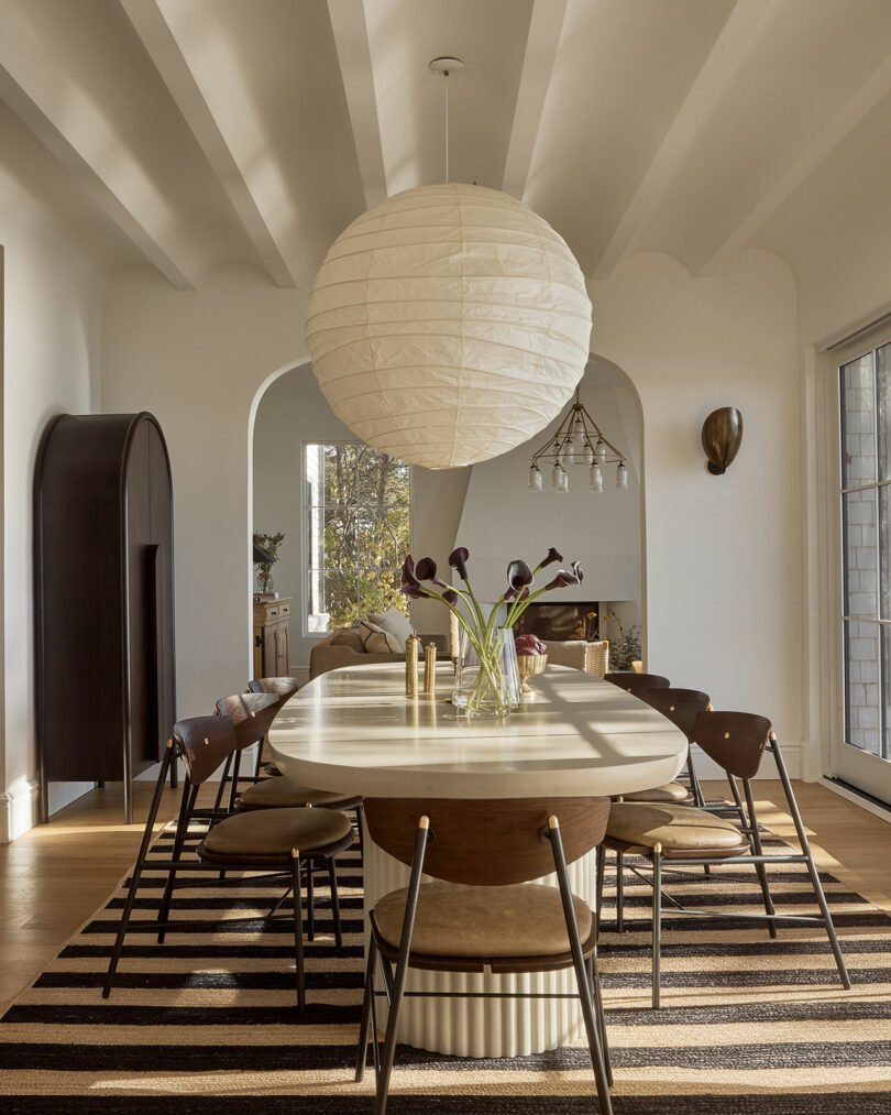A modern dining room by Sam Sacks features a long oval table, wooden chairs, a striped rug, a large paper lantern pendant light, and a vase of dark flowers on the table.
