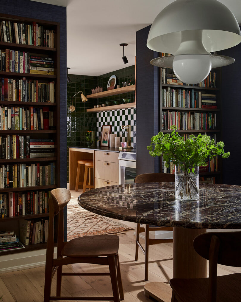 A dining area with a round marble table, four wooden chairs, a glass vase with greenery, bookshelves, and a kitchen with Sam Sacks-inspired green tiled walls in the background.