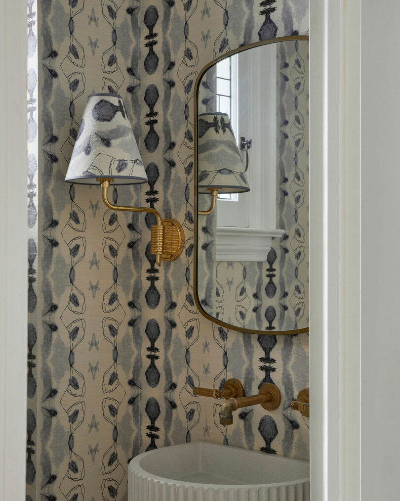 A small bathroom by Sam Sacks features patterned wallpaper, a scalloped white sink, gold faucet, oval mirror, and a wall sconce with a patterned shade.