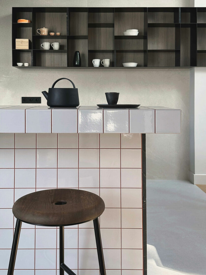 A modern kitchen features a tiled counter with a black teapot, cup, and saucer, a wooden stool, and open shelving with minimalist dishware in the background.
