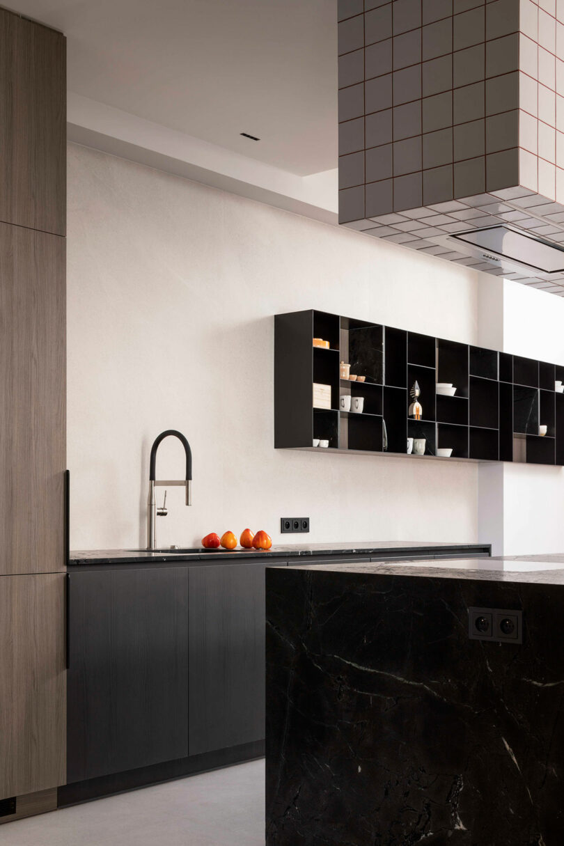 Minimalist kitchen with dark cabinets, a black marble island, open shelving with decor items, a modern faucet, and several oranges on the countertop.