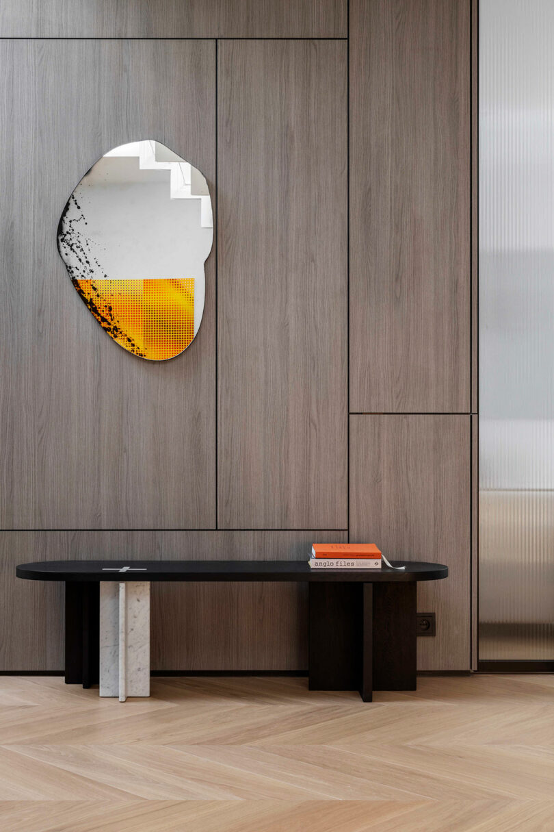 A modern bench with a marble leg stands against a wood-paneled wall, beneath an abstract mirror and next to a stack of orange and white books.