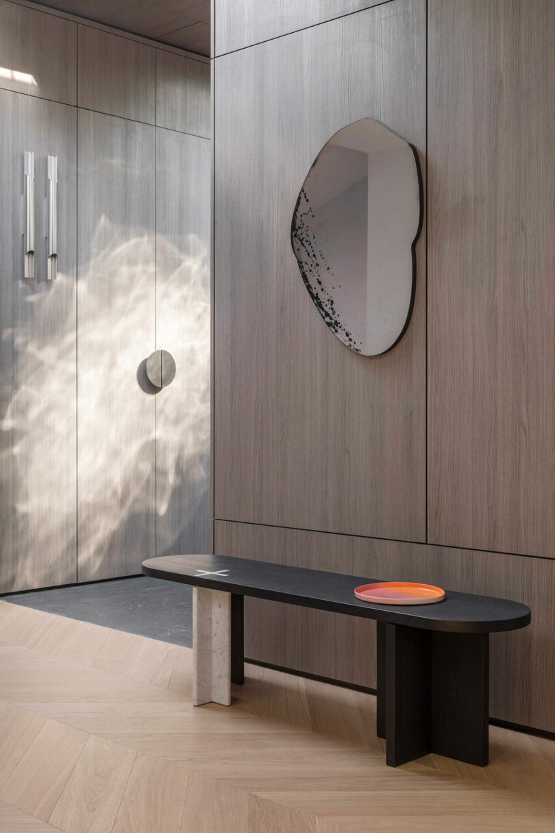 Modern minimalist entryway with light wood paneling, abstract wall mirror, black bench with mixed legs, and two colored trays on the bench. Sunlight casts patterns on the wall.