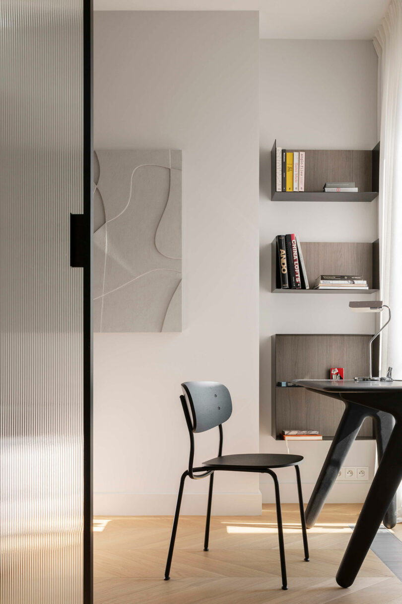 Minimalist home office with a black chair, dark desk, wall-mounted shelves holding books, and abstract wall art in a light-filled room.