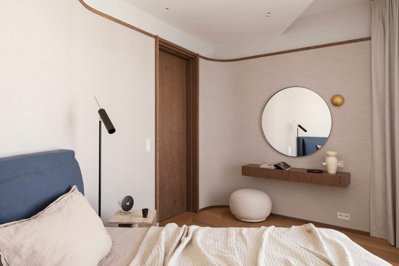 Minimalist bedroom with a blue upholstered bed, beige bedding, round mirror above a floating wooden shelf, and neutral-colored walls and furniture.