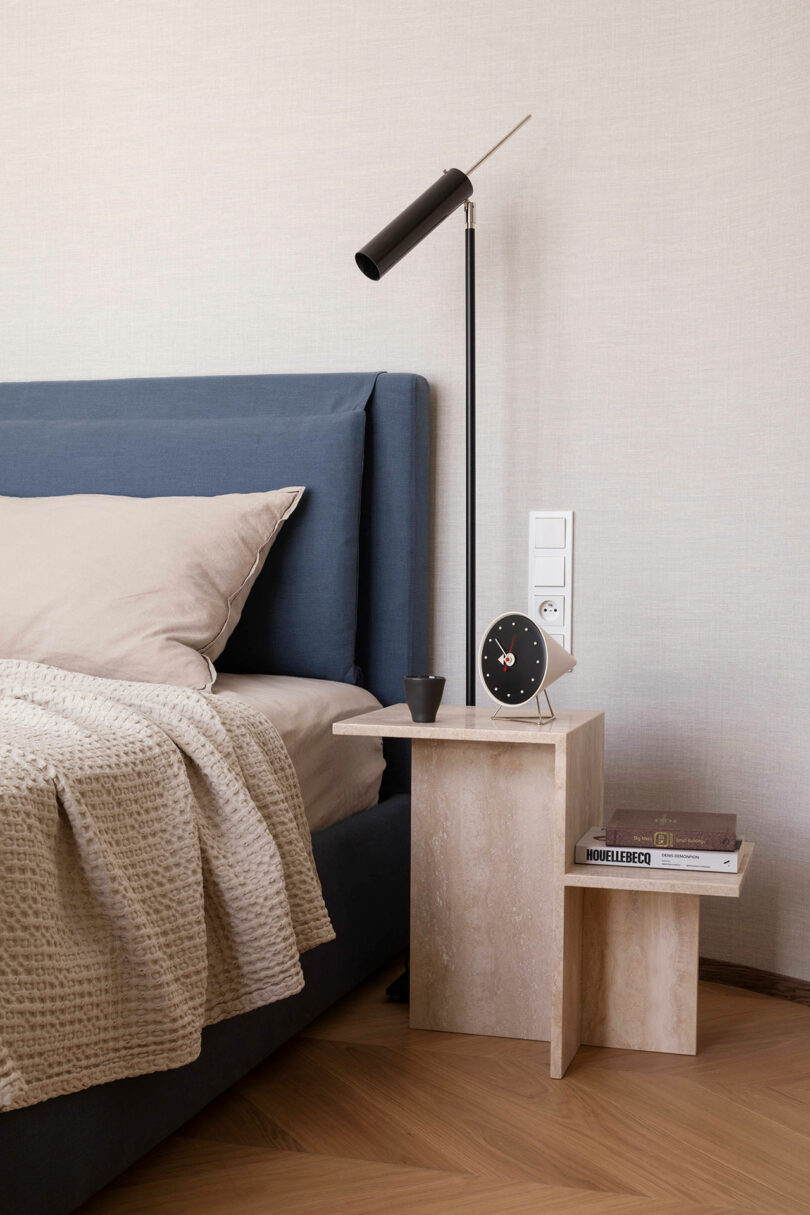 A modern bedside table with a clock, a small cup, a stack of books, and a black lamp beside a blue upholstered bed with beige bedding.
