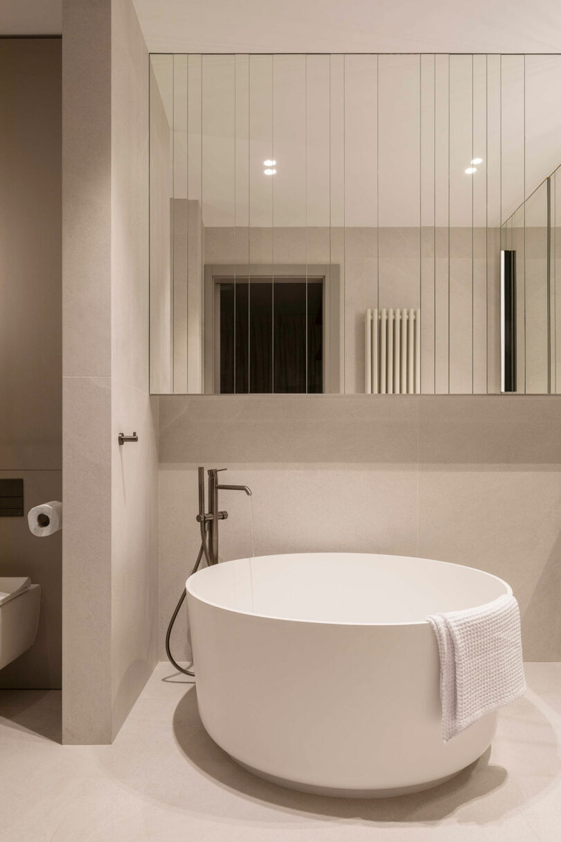 Minimalist bathroom with a round freestanding bathtub, wall-mounted faucet, large mirror with vertical lines, and a white towel draped over the tub edge.