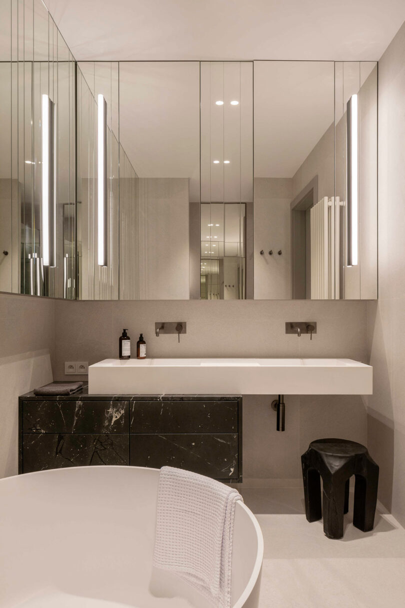 Modern bathroom with a large mirror, wall-mounted sink, black vanity, minimalist lighting, a white tub with a towel, and a black stool.