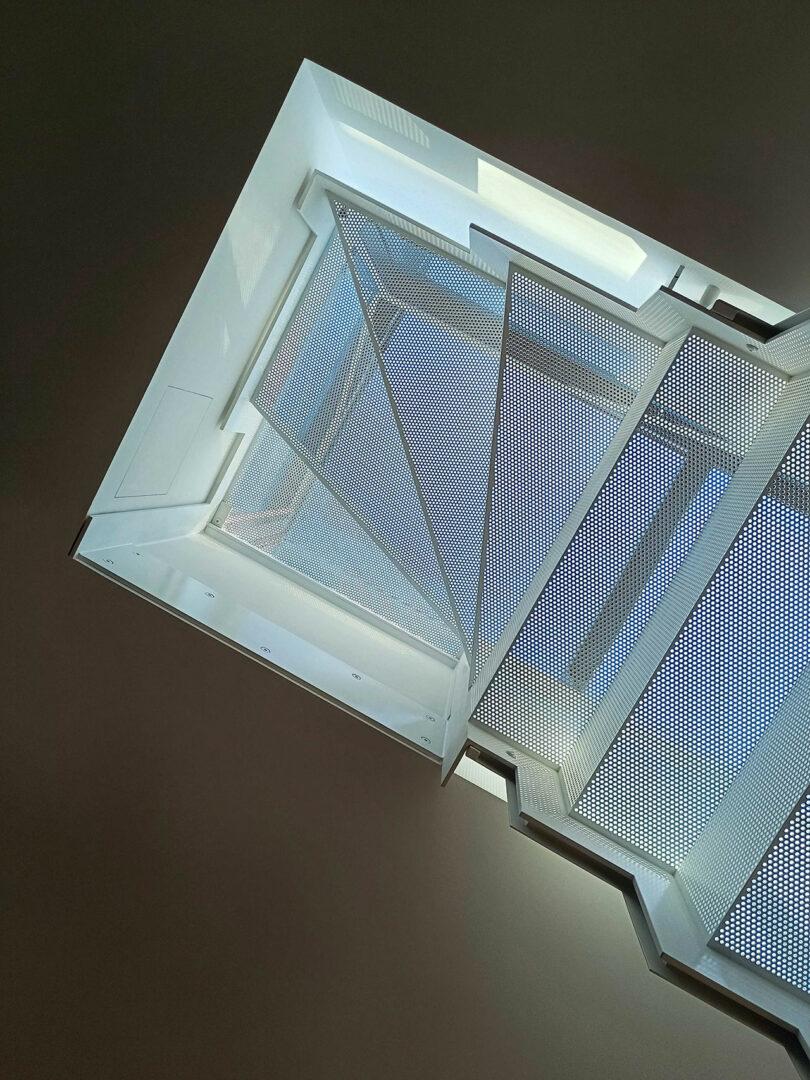 View looking up a modern, white metal staircase with perforated steps and geometric lines against a dark background.