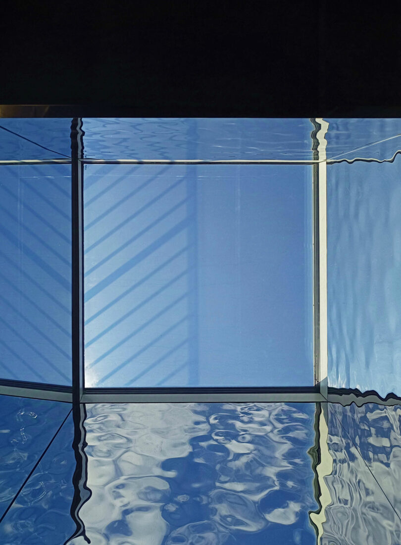 View of rippling water above a rectangular glass ceiling with shadows from window frames cast on a bright blue sky.