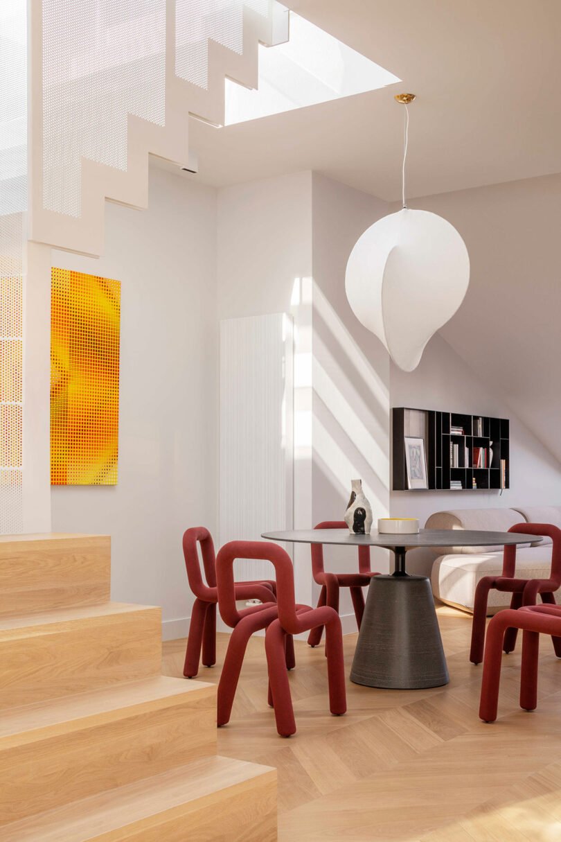 Modern dining area with red sculptural chairs, a round dark table, a large white pendant light, wooden stairs, abstract yellow wall art, and natural light streaming through a skylight.