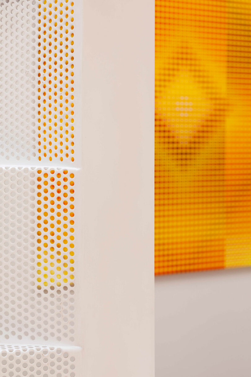Close-up of a white perforated metal panel on the left, with an out-of-focus orange and yellow patterned surface in the background on the right.