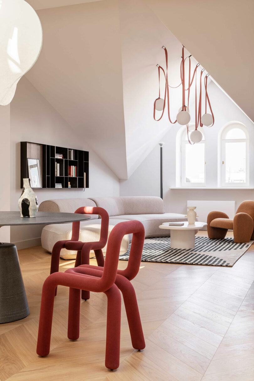 Modern living room with unique red chairs, a light grey sofa, round tables, geometric decor, and pendant lights hanging from a sloped ceiling. Natural light enters through arched windows.
