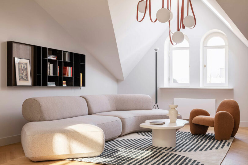 Modern living room with a beige sectional sofa, orange armchair, white coffee table, patterned rug, and wall-mounted bookshelf under slanted ceiling with large arched windows.